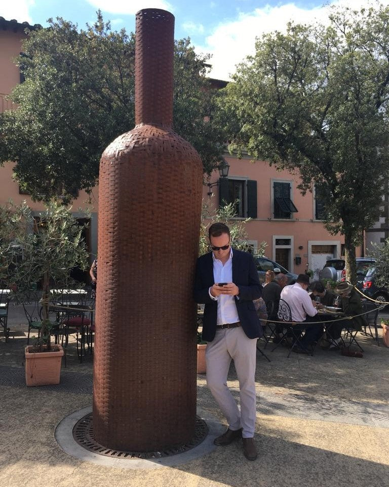 Andrew Catt beside a giant wine bottle sculpture in a Tuscan square